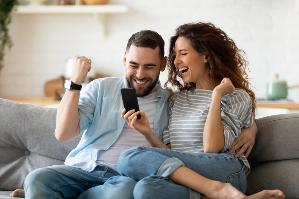 Excited overjoyed couple resting on couch holding smart phone celebrating