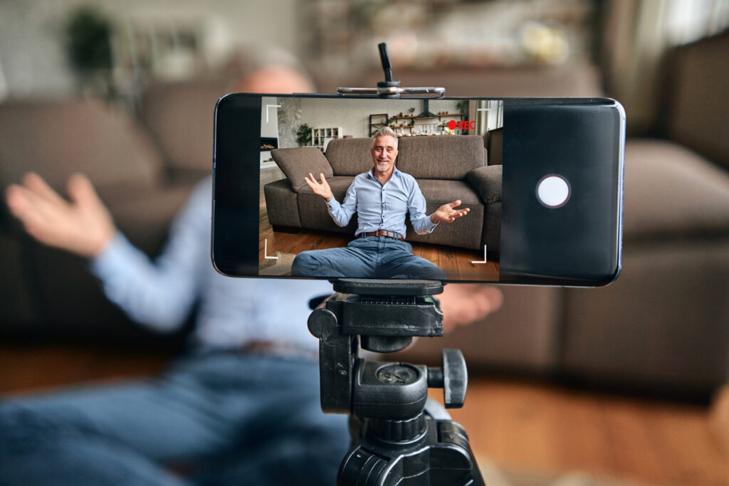 Happy adult man shooting video content for his vlog while sitting on the floor in living room at home. Blogging and lifestyle concept